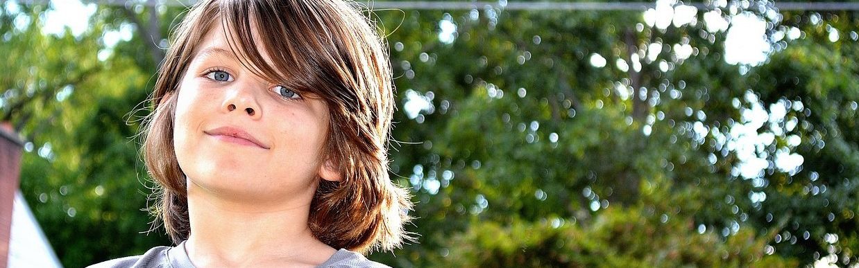 Junge mit schulterlangen Haaren blickt selbstbewusst in die Kamera, im Hintergrund grüne Bäume