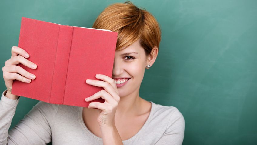 Frau lächelt hinter rotem Buch vor grüner Tafel