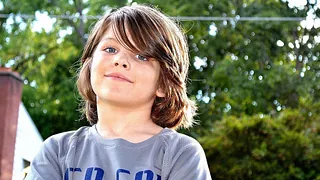 Junge mit langen Haaren blickt selbstbewusst in die Kamera, Bäume im Hintergrund