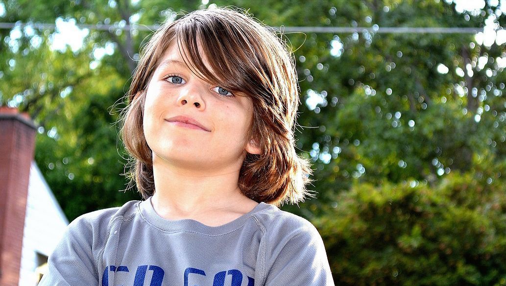 Junge mit langen Haaren blickt selbstbewusst in die Kamera, Bäume im Hintergrund